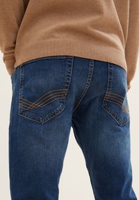 Hombre que lleva jeans de mezclilla azules con bolsillos traseros cosidos y un suéter marrón con canalé, mostrado desde la cintura hacia abajo, vista trasera.