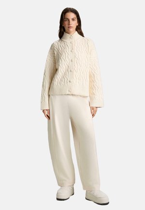 Donna che indossa un cardigan crema a trecce con bottoni, pantaloni crema a gamba larga e sneakers abbinate su uno sfondo chiaro e uniforme.