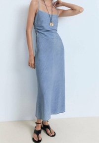 Robe maxi en denim avec des bretelles fines, teinte bleu clair, forme épurée et texture lisse. Assortie à des chaussures de style sandales noires.