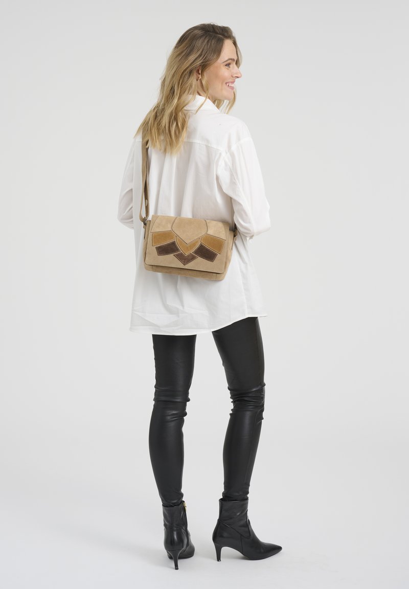 Femme portant une chemise blanche, un pantalon en cuir noir, des bottes à talons noires et un sac bandoulière beige avec des patchs géométriques, souriant en étant de profil.