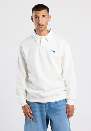 Witte longsleeve polo sweatshirt met een kraag, voorzien van een blauw "NYC" logo op de linkerkant van de borst en een relaxte pasvorm. Spijkerbroeken zichtbaar eronder.