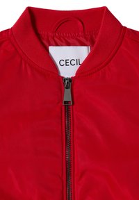 Veste bomber rouge avec col côtelé, fermeture éclair en métal noir et étiquette blanche montrant une partie du nom de la marque "CECIL".
