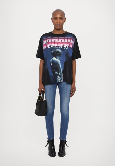 Svart grafisk T-skjorte med et trykk av en blå og hvit statue og tekst. Kombinert med blå skinny jeans og svarte ankelstøvler, mens hun holder en svart håndveske.