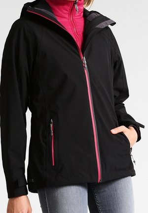 Frau trägt eine schwarze Jacke mit pinkfarbenen Reißverschlussdetails über einem knallpinken Oberteil, die Hand in der Tasche, kombiniert mit hellblauen Jeans.
