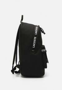 Black backpack with adjustable straps, textured fabric, a side pocket, and prominent "LACOSTE" branding on the strap. Closed zipper.