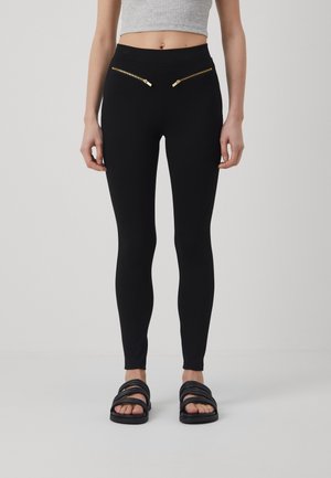 Leggings noirs à taille haute, dotés de deux poches zippées avec quincaillerie dorée à l'avant. Texture lisse et coupe près du corps.