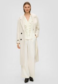 Cremefarbener, übergroßer Mantel aus strukturiertem Stoff, schwarze Knöpfe; geschichtete, gefaltete weiße Bluse, weite Hose, schwarze Stiefeletten.