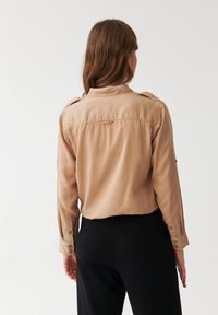 Beige shirt met knoopmanchetten en schouderechterstukken, gemaakt van een gladde stof. Zwarte broek is zichtbaar, met een rechte snit en achterzakken.