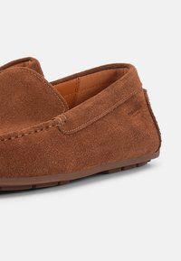 Καφέ σουέτ μοκασίνι ανδρικό με ραφές και καουτσούκ σόλα, εμφανιζόμενο σε κοντινή λήψη σε λευκό φόντο.