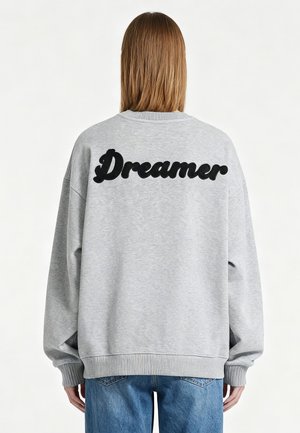 Personne aux cheveux lisses châtain clair portant un sweat-shirt gris avec « Dreamer » écrit en noir dans le dos et un jean bleu.