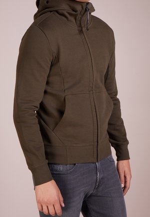 Homme portant un sweat à capuche zippé vert olive avec poches avant et poignets côtelés, associé à un jean gris foncé.