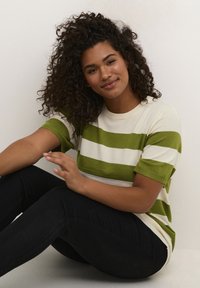Grøn og creme farvet stribet striktrøje med korte ærmer, parret med sorte skinny jeans. Modellen sidder mod en hvid baggrund.