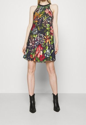 Femme portant une robe plissée sans manches à motif floral sur fond noir et des bottines noires, debout contre un mur clair uni.