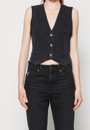 Femme portant un gilet sans manches en denim noir boutonné et un jean noir taille haute, debout devant un fond blanc uni.