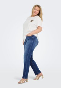 White short-sleeve t-shirt with a leopard-patterned heart design, paired with dark blue jeans and beige block-heeled sandals.