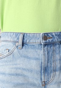 Jeans denim berwarna biru muda dengan pinggang tinggi, jahitan oranye, dan penutupan kancing di depan. Dipadukan dengan atasan lengan pendek berwarna hijau.