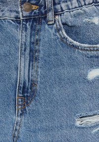 Gros plan sur un jean en denim bleu avec des coutures marron, un bouton métallique, et des trous effilochés usés près de la poche et de la cuisse.