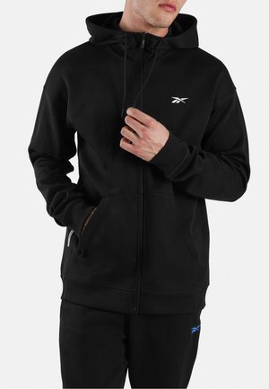 Hombre con sudadera negra con cremallera y pantalones a juego con logos blancos y azules de Reebok, ajustando el cordón de la sudadera con una mano.