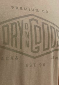 Kaki pamučna majica s velikom prigušenom grafikom koja prikazuje natpise "DRY DNM" i "EST. 90." u tamnozelenom tisku. Jednostavna pozadinska tekstura.
