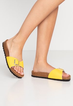 Gule slidesandaler med en blank finish, der har en spænde detalje, korksål og tekstureret sort gummisål.
