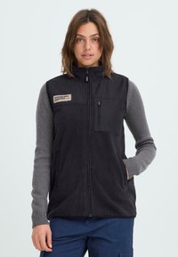 Schwarze Fleece-Weste mit hohem Kragen, Frontreißverschluss und linker Brusttasche. Mit Patch-Details und seitlichen Taschen, über grauen Pullover-Ärmeln getragen.
