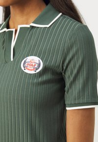 Zelený ribbovaný polo tričko s bielymi akcentmi a golierom. Obsahuje kruhovú nášivku s textom „GOLF Club“ a dizajnom vavrínu. Krátke rukávy.