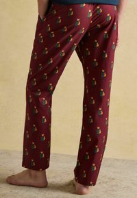 Cilvēks, kurš valkā bordo bikses ar atkārtotu lapsas tēlu rakstu, stāv basām kājām uz teksturētas grīdas pret vienkāršu bēškrāsas fona.