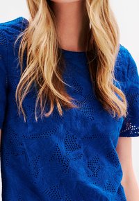 Haut en dentelle bleu avec un motif floral texturé, manches courtes et encolure ronde. Le matériau présente des découpes et une finition lisse.