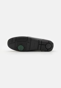 Sola di scarpa in gomma nera con texture a motivo, caratterizzata da un accento circolare verde e da un logo del marchio inciso. La forma è piatta e rettangolare.