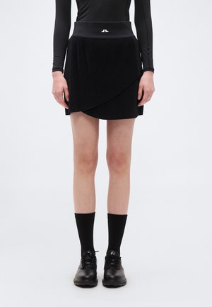 Kvinde iført en sort plisseret slå-om-nederdel, sort bluse med lange ærmer, sorte mellemhøje sokker og sorte snøresko på en hvid baggrund.