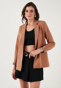 Smeđa blazer jakna s prilagođenim dizajnom, dugim rukavima i leopard uzorkom na manžetama. Nosena preko crnog crop topa i crne suknje.