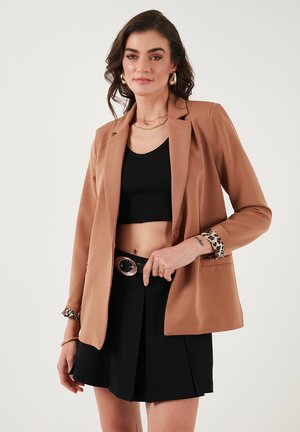 Blazer marron avec un design ajusté, manches longues et accents en imprimé léopard aux poignets. Porté sur un crop top noir et une jupe noire.