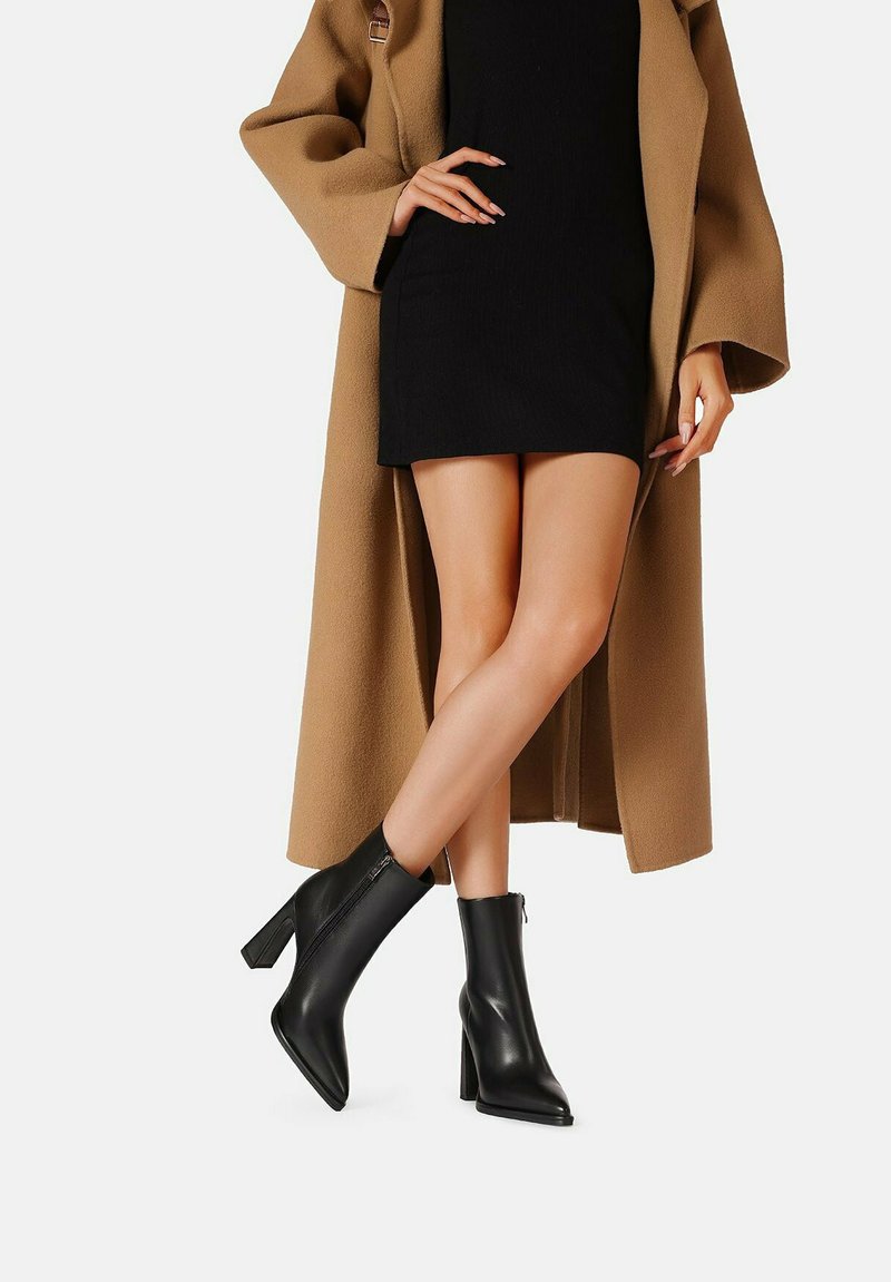 Mujer con abrigo color camel, vestido corto negro y botines negros de tacón, de pie con las piernas cruzadas sobre fondo blanco.