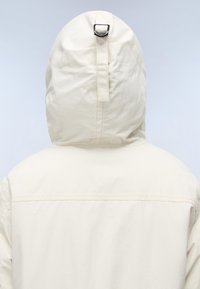 Lys beige jakke med hette, med en vertikal søm langs ryggen, og en svart spenne detalj øverst på hetten.