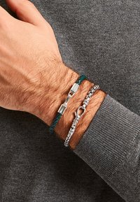 Zwei Armbänder an einem Handgelenk: ein grünes geflochtenes Lederarmband mit einem silbernen Verschluss und ein glänzendes silbernes Kettenarmband mit einem Karabinerverschluss.