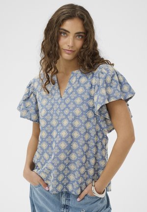 Žena s vlnitými hnedými vlasmi, oblečená v modrej blúzke s vzorom a rozviatymi rukávmi, svetlomodré džínsy, ruky v kapsách, neutrálny pozadie.