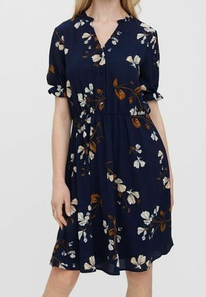 Kobieta w granatowej sukience do kolan z białymi i brązowymi kwiatowymi wzorami, krótkimi bufkowatymi rękawami i lekko marszczoną talią.