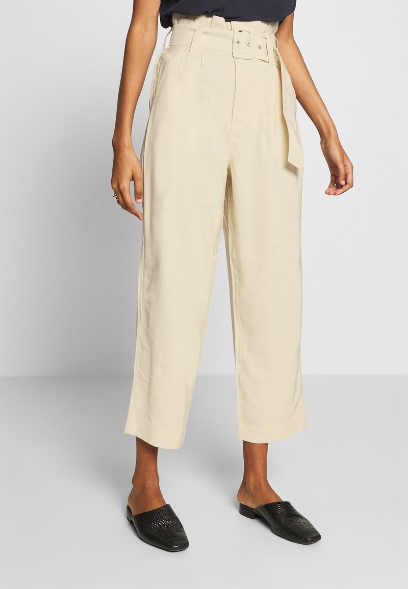 Kvinna i högmidjade beiga vida capribyxor med bälte, svarta slip-on skor, som står mot en enfärgad bakgrund.