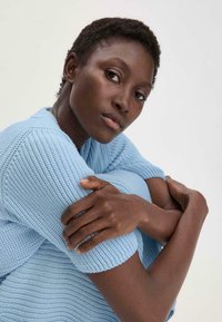 Lichtblauwe gebreide trui met korte mouwen, gestructureerd oppervlak en geribde manchetten. Model heeft kort haar en heeft haar armen gekruist.