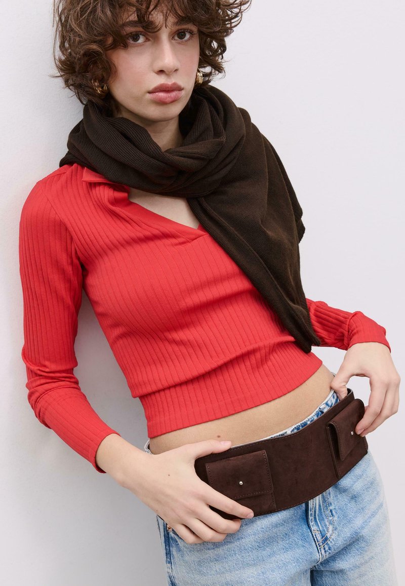 Rdeča rebrasta majica z dolgimi rokavi, kombinirana s temno rjavo ruto. Obleka vključuje širok rjav pas z dvema žepoma, nošen preko svetlo modrih kavbojk.