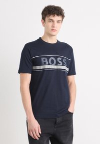 Tamsiai mėlynas medvilninis marškinėliai su ryškiu pilku "BOSS" logotipu krūtinėje, trumpomis rankovėmis ir apvalia kaklo iškirpte.