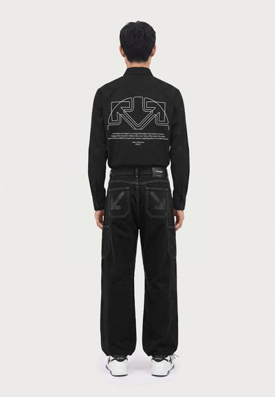 Homme de dos portant un t-shirt noir avec un motif de flèche blanche et un texte, pantalon noir avec des imprimés de flèches sur les poches arrière, et des baskets blanches.