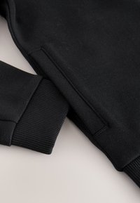 Schwarzer Pullover mit strukturiertem Stoff, gerippten Bündchen und glatter Oberfläche. Das Design ist schlicht, ohne sichtbare Logos oder Muster.