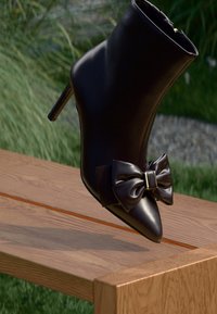 Bottine noire en cuir avec un bout pointu, présentant un grand nœud accentué à l'avant et un talon slim haut, posée sur une surface en bois.