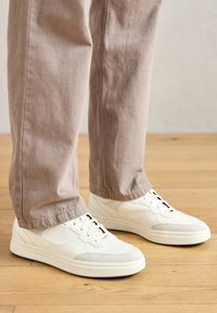 White sneakers with textured leather and smooth accents, featuring a rounded shape, lace-up design, and black eyelets, paired with beige pants.