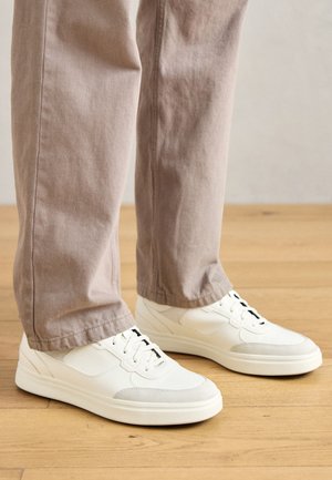 Biele tenisky s textúrovanou kožou a hladkými akcentami, s okrúhlym tvarom, šnurovaním a čiernymi okuliarmi, spárované s béžovými nohavicami.