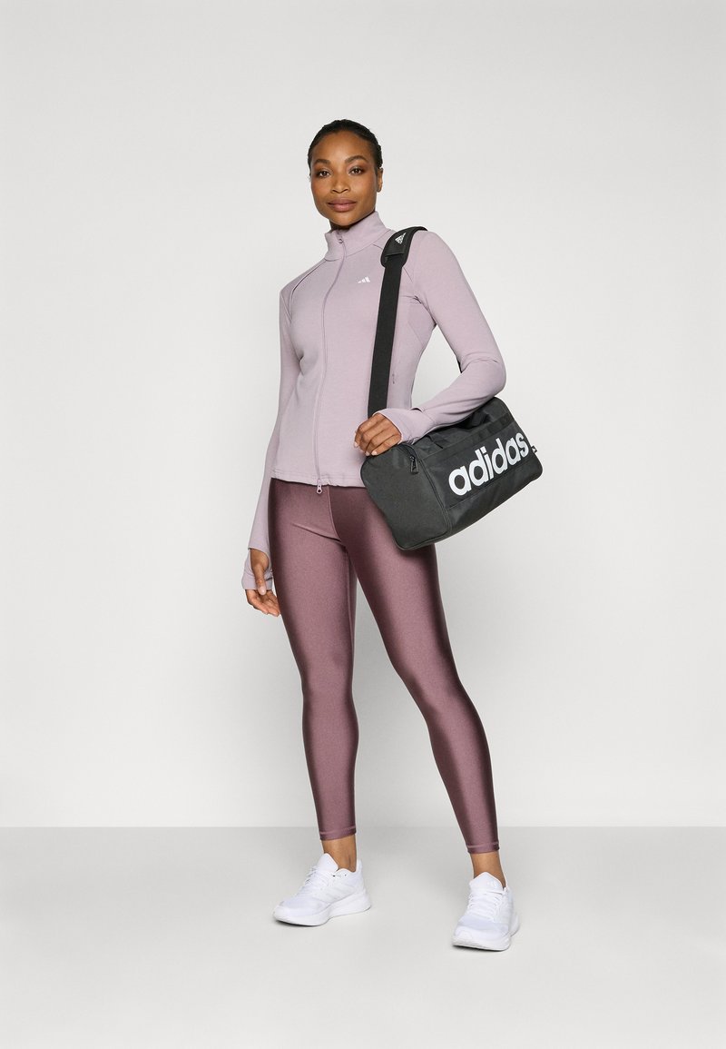 Taillierte lavendelfarbene Longsleeve-Jacke, metallisch pinke Leggings und weiße Sneakers. Schwarze Adidas-Tasche mit weißem Logo. Glatte Texturen in der gesamten Ausstattung.