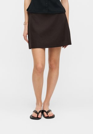 Femme portant une jupe noire jusqu'au genou, un haut noir boutonné, des tongs noires, et un bracelet porte-bonheur au poignet gauche.