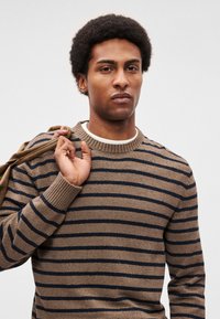 Πουλόβερ με ρίγες σε καφέ και ναυτικό μπλε, πλεκτή υφή, στρογγυλή λαιμόκοψη, ριμπ μανσέτες, φορεμένο με μπεζ μπουφάν περασμένο πάνω από τον ώμο.