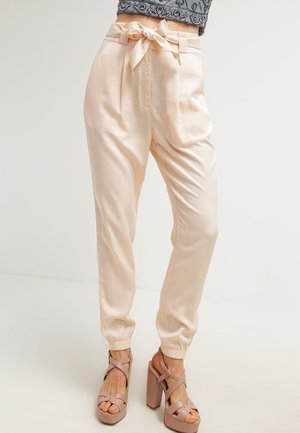 Femme portant un pantalon beige taille haute avec une ceinture nouée en forme de nœud et des poignets élastiques, associé à des sandales compensées nude à talons épais.